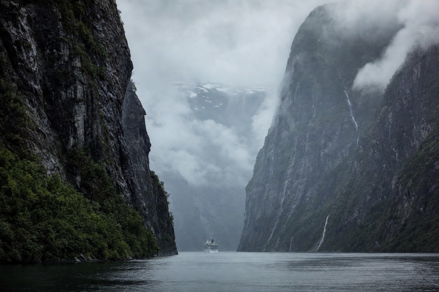 Very narrow fjord walled by dark cliffs with a small cruise boat dwarfed between them under low mist