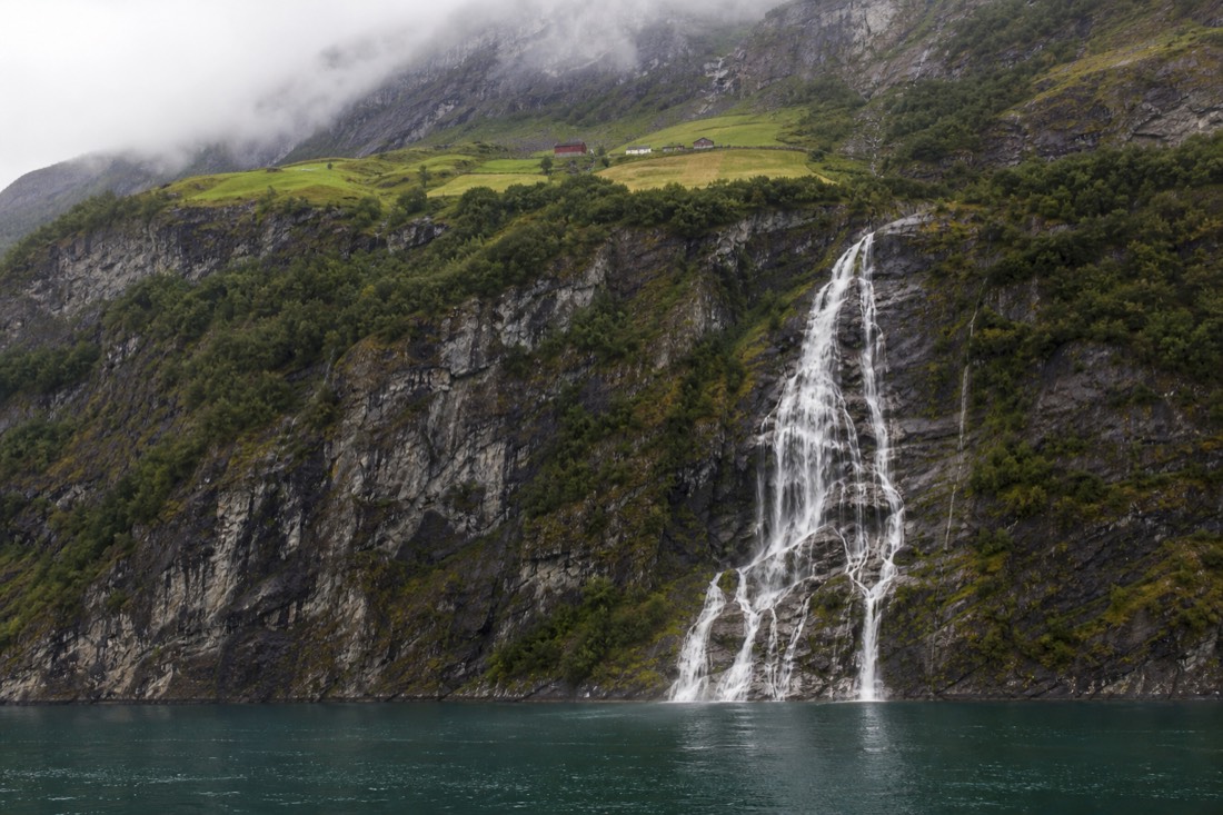 A multi-strand waterfall spilling down a sheer rock face into a narrow fjord with a small ledge farm above