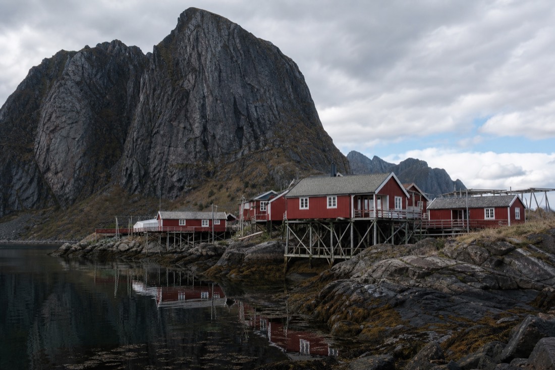 Red wooden fishing cabins on stilts at the foot of steep dark granite peaks reflected in still Arctic water
