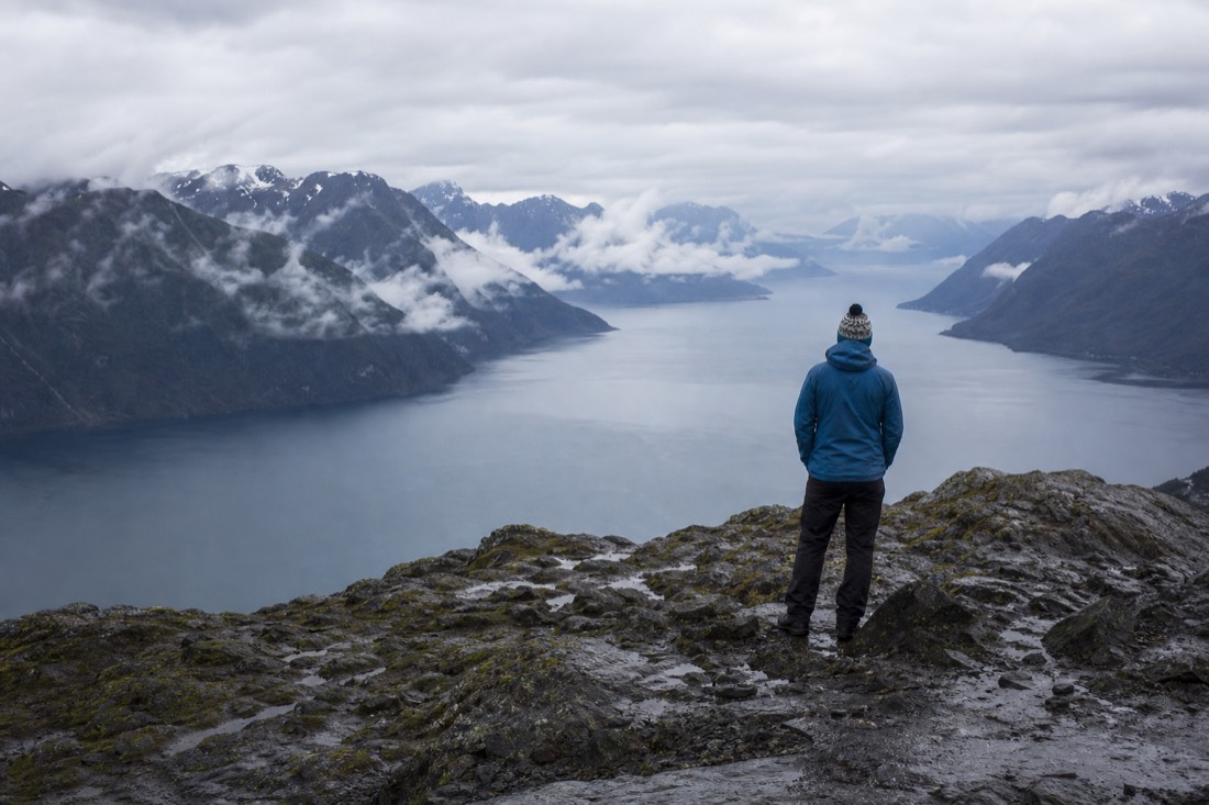 Hiker in a shell jacket and wool hat standing on a rocky ridge above a cloudy fjord