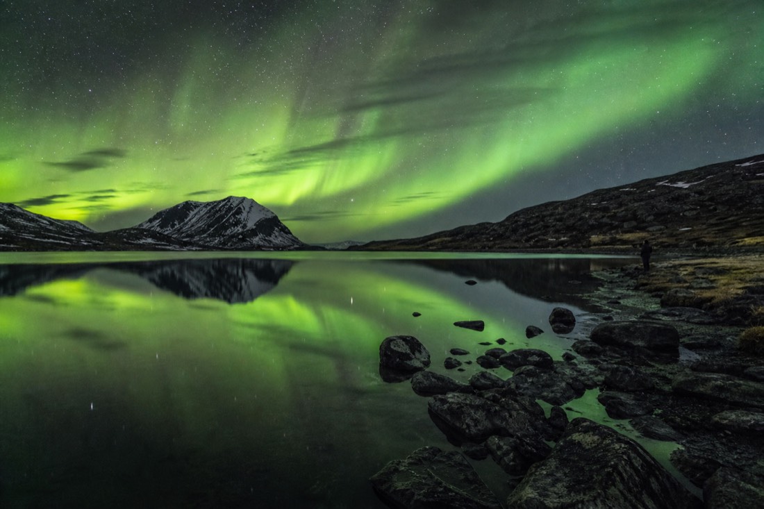 Aurora reflected in still dark water away from town lights with a lone figure watching from the shore