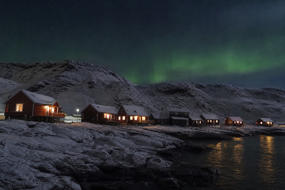 Row of snow-covered Arctic cabins at night with lit windows and faint green light above the ridge