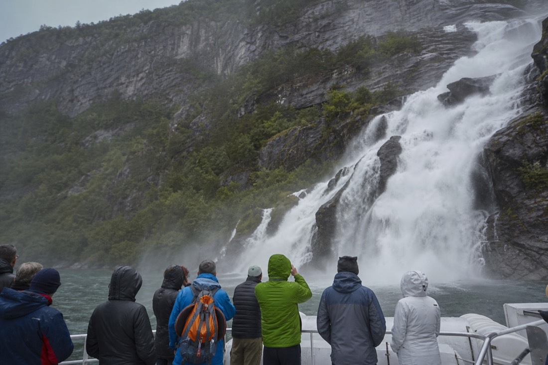Passengers on an open deck as a fjord boat approaches a roaring waterfall in Osterfjord