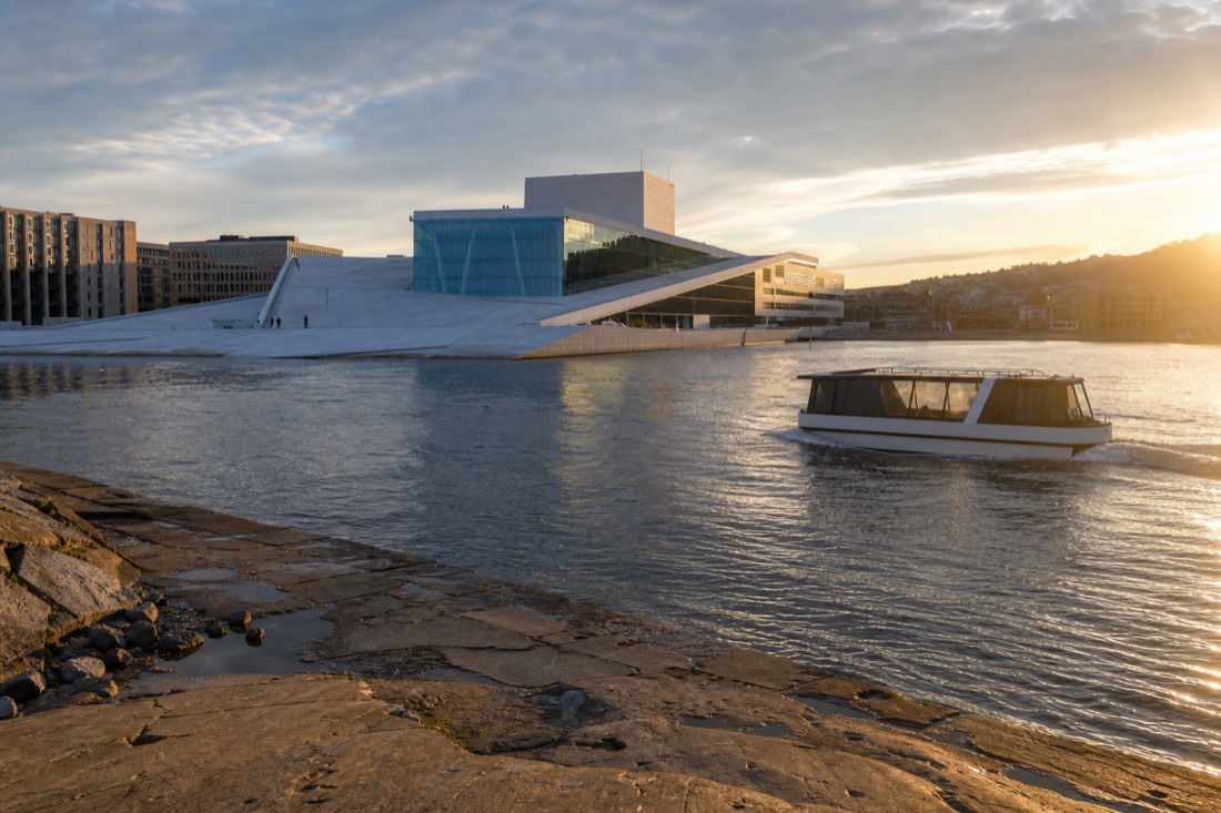 A silent electric sightseeing boat gliding past the white marble Oslo Opera House at golden hour