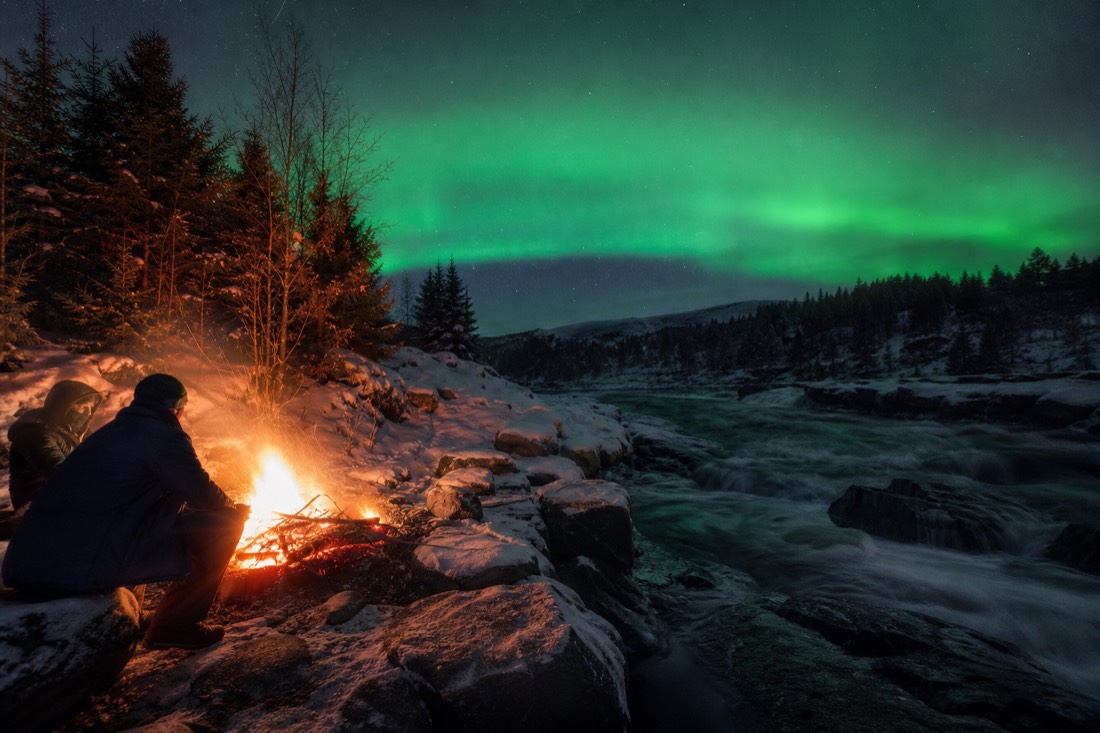 Travelers warming their hands over a bonfire in the snow while the aurora flickers above the treeline
