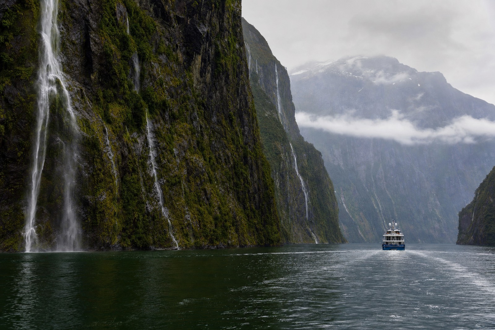 A sightseeing boat sailing calm fjord water between mossy cliff walls streaked with waterfalls