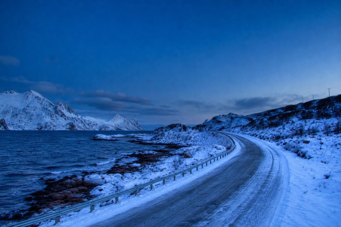 Snow-covered Arctic road curving between low mountains under a deep blue winter twilight