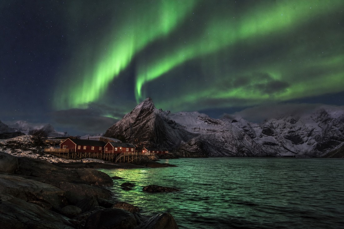 Green aurora over snow-covered peaks and a dark Arctic bay with fishing cabins below
