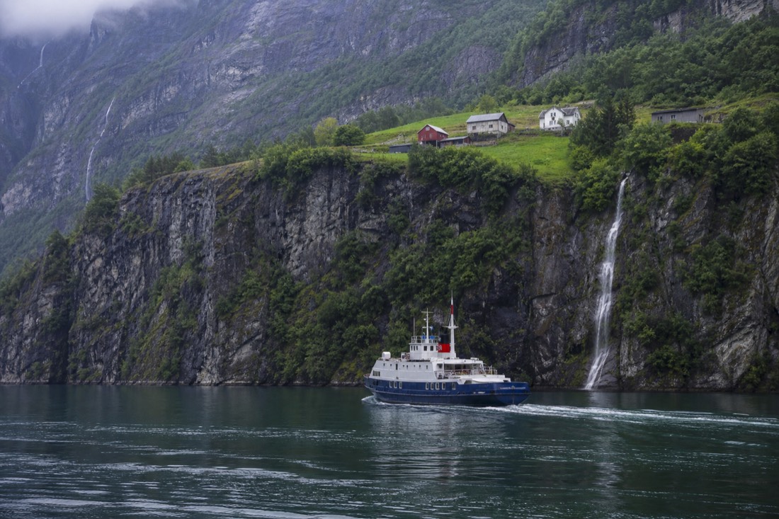 Narrow steep-sided Norwegian fjord with a small passenger ferry passing beneath cliffs and farm buildings