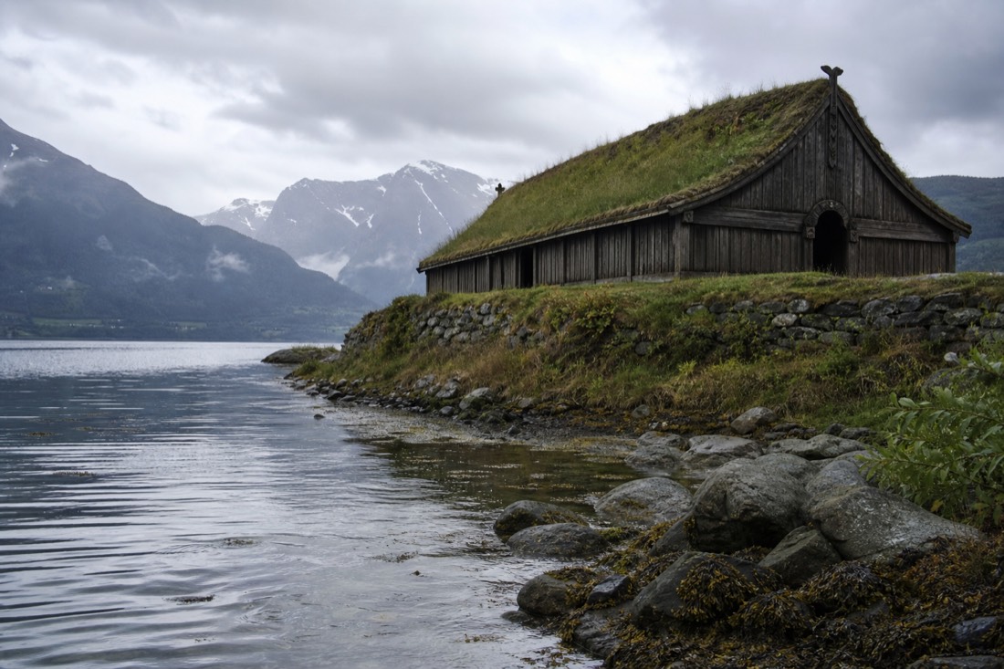 Timber Viking longhouse with turf roof beside a fjord shoreline under overcast Norwegian sky