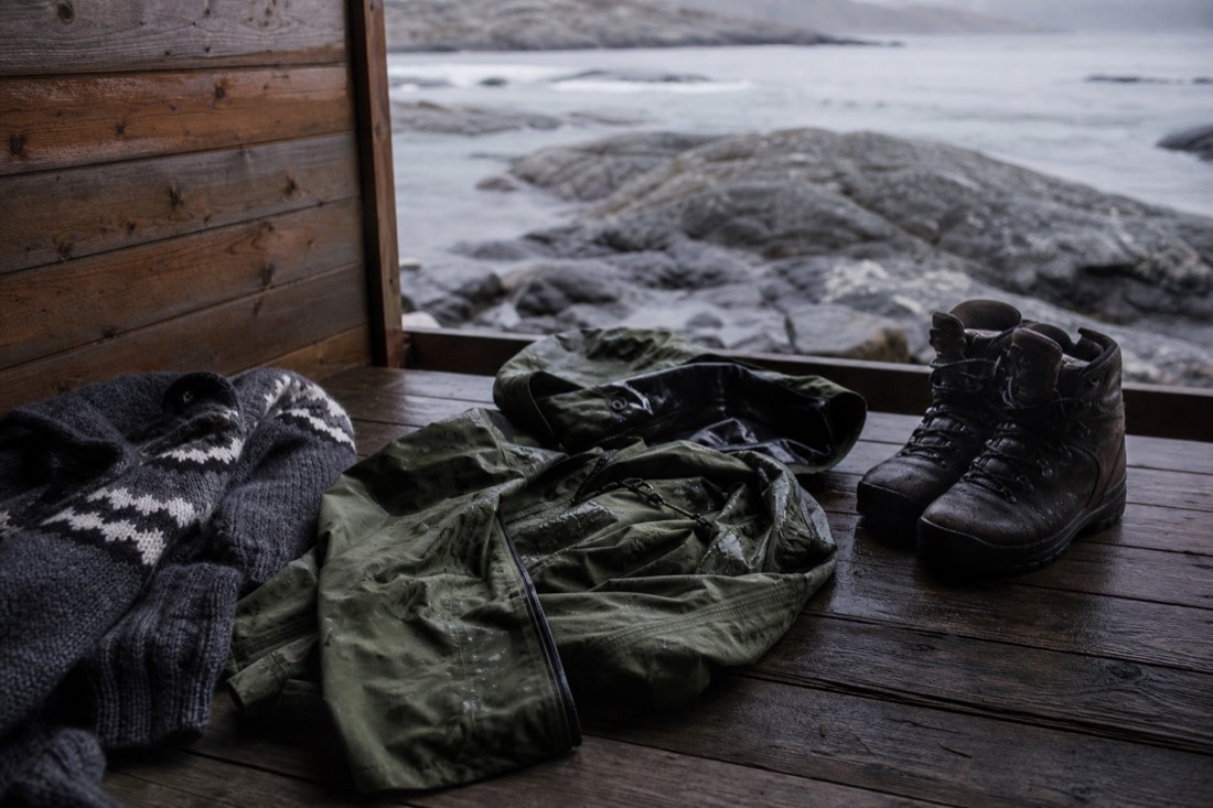 Wool sweaters, waterproof shell jacket and winter boots laid out on a wooden cabin floor