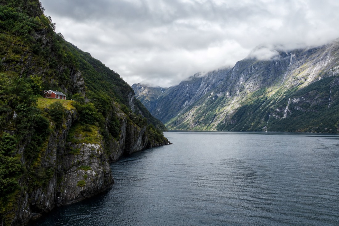Steep green fjord walls dropping into deep blue water with a small red farmhouse on a ledge