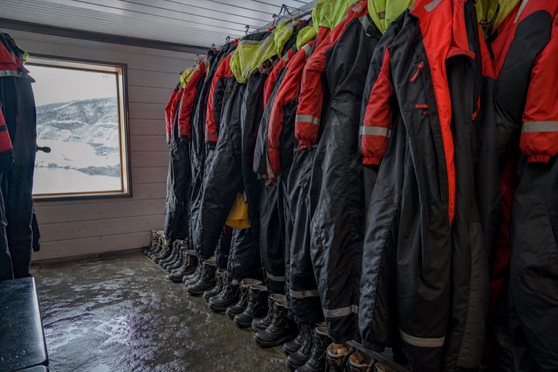 Row of thermal suits and boots hanging in a heated tour cabin with snow visible through a small window