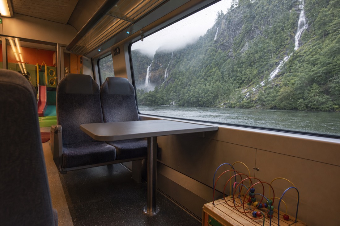 Interior of a Norwegian train carriage with a small play area, seats facing a table and forest passing outside