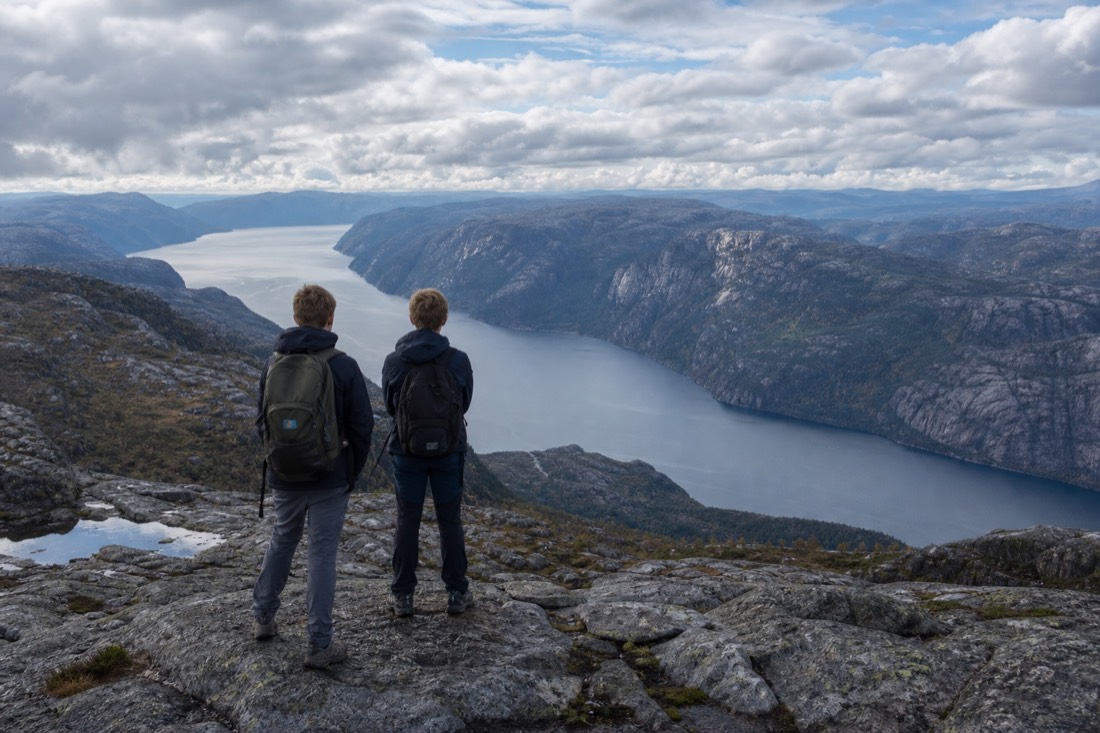 Two teenagers in hiking gear on a rocky ridge looking down at a long fjord under scattered cloud
