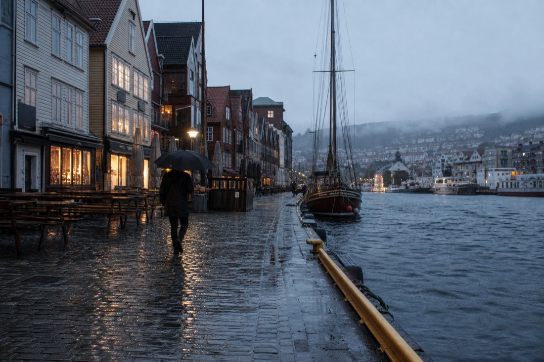 Rain-slicked cobbles reflecting shopfront lights with a person under a black umbrella walking past moored boats