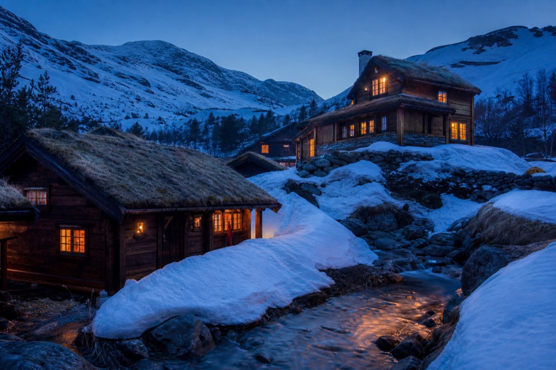 Cluster of turf-roofed timber ski cabins lit from within at blue hour with deep snow banks between them