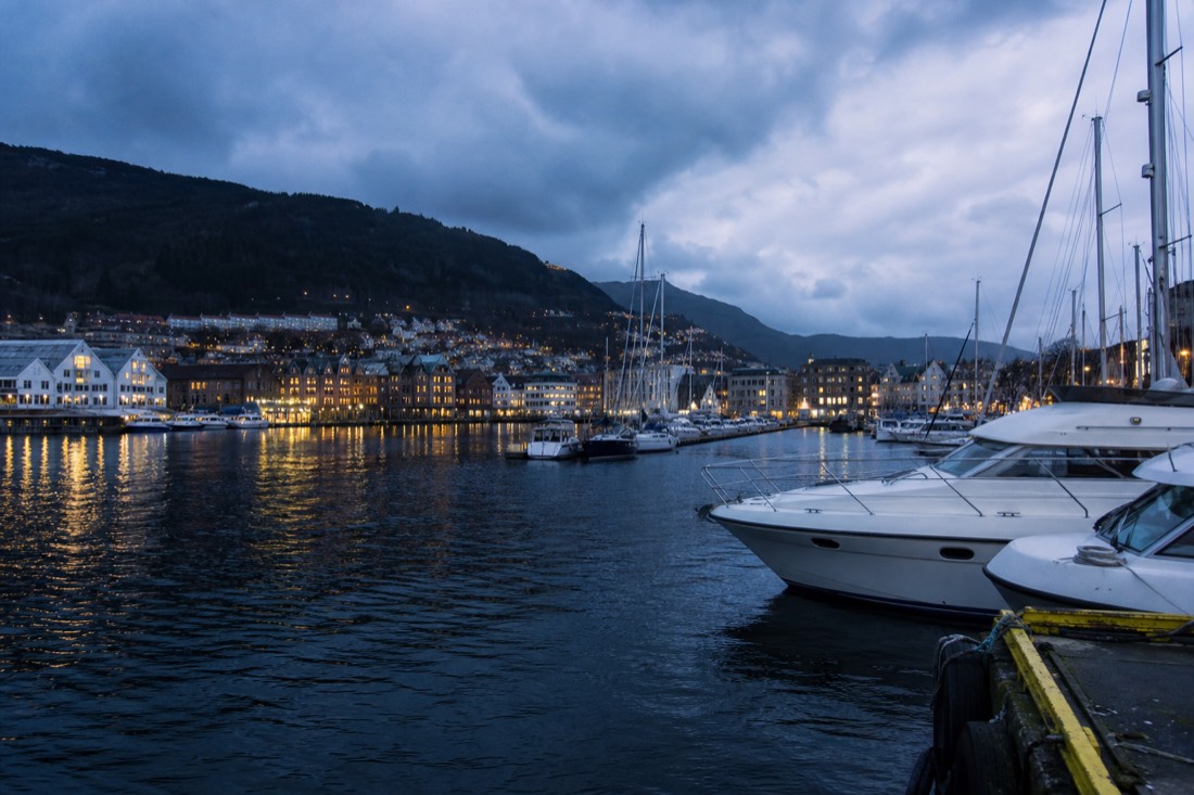 Bergen harbour at dusk with moored boats, lit quayside windows and mountains dark behind the town