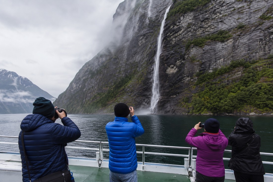 Passengers on an open boat deck photographing sheer cliffs and a waterfall in a narrow Norwegian fjord