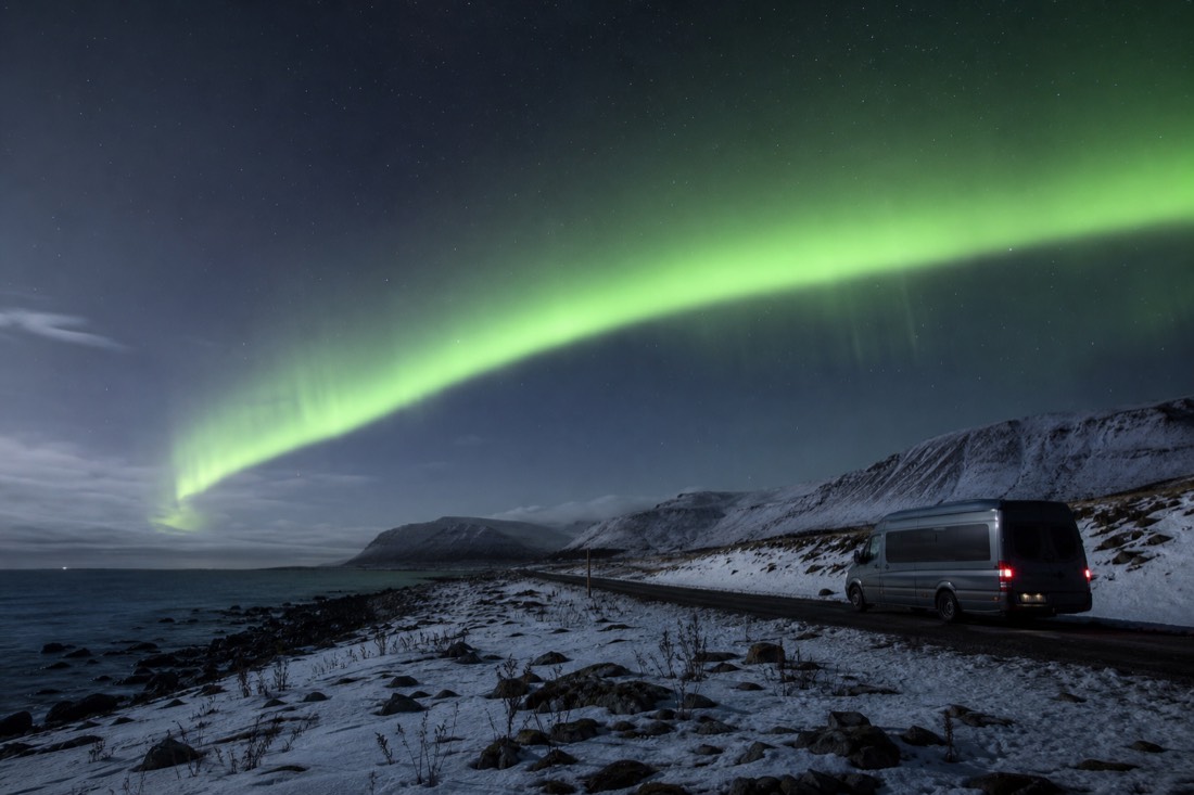 Green aurora arc over a snow-covered shoreline with dark mountains and a parked minibus on a coastal road