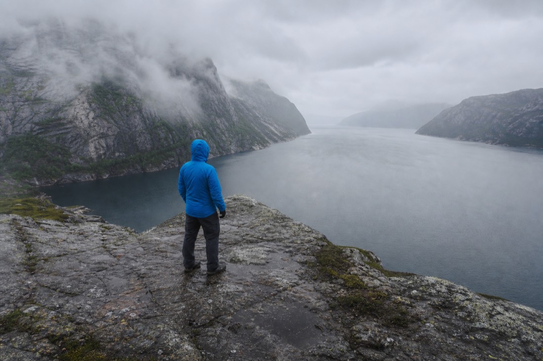 Hiker in a bright waterproof shell jacket standing on wet rock above a grey fjord under low cloud